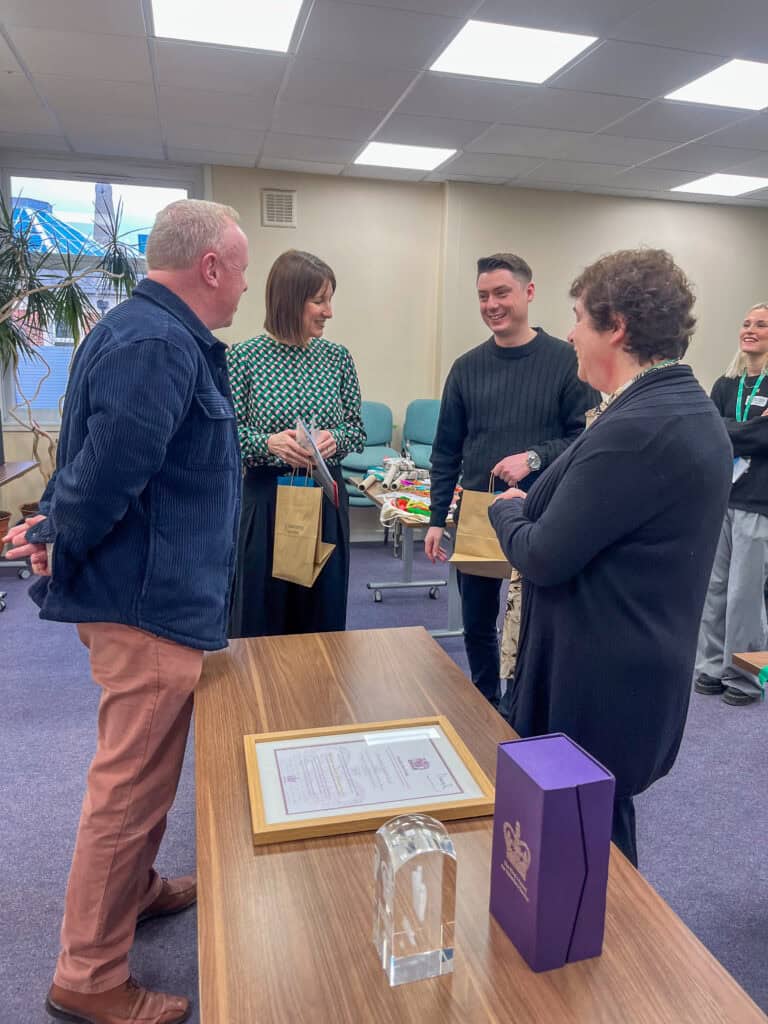 A group of four people stand together in a circle laughing. Two hold brown paper bags with aprons in. On the table next to them is an award and certificate.