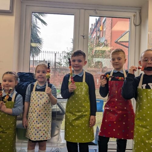 An image of five children wearing aprons, happily holding skewers of food they prepared during a cooking activity at Flourishing Families.